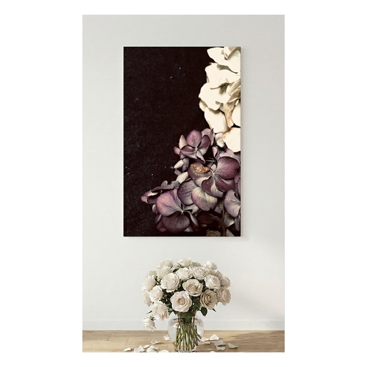 A vertical canvas wall art print showing a close-up floral composition with cream and mauve-purple flowers on a dark background, displayed above a vase of matching white flowers on a light wood floor against a white wall.