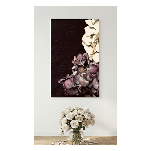 A vertical canvas wall art print showing a close-up floral composition with cream and mauve-purple flowers on a dark background, displayed above a vase of matching white flowers on a light wood floor against a white wall.