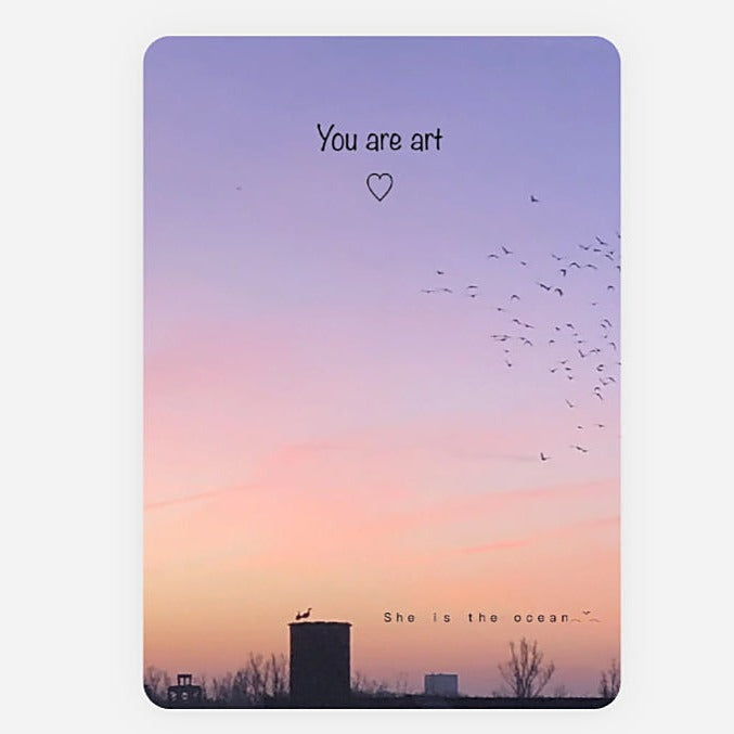 A vertical greeting card art print or card featuring a pastel sunset sky with the quote 'You are art' and a small heart near the top, and 'She is the ocean' with silhouetted buildings and birds near the bottom.