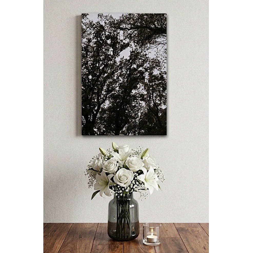 A vertical black and white canvas wall art print depicting tree branches, displayed on a light gray wall above a wooden table with a vase of white flowers and a small candle.