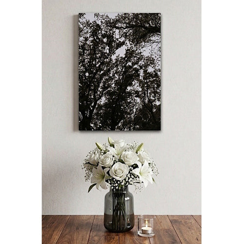 A vertical black and white canvas wall art print depicting tree branches, displayed on a light gray wall above a wooden table with a vase of white flowers and a small candle.