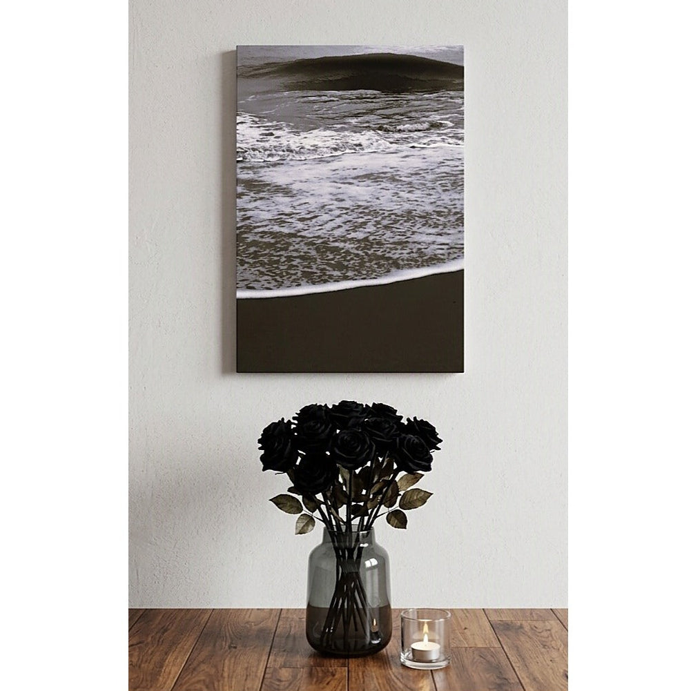 A vertical ocean artwork canvas wall art print depicting a dark beach scene with ocean waves, displayed on a light gray wall above a wooden table with black roses in a vase and a small candle.