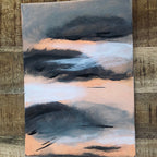 An abstract emotional art print landscape painting on canvas featuring dark charcoal and slate blue clouds over a soft peach and light blue sky, displayed on a rustic wooden background.