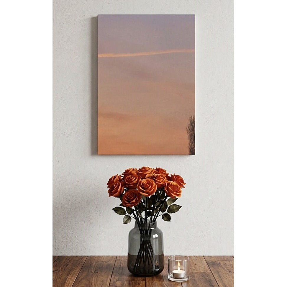 A vertical canvas wall art print depicting a soft sunset sky in peach, orange, and muted lavender tones, styled in a home setting above a wooden table with a vase of orange roses and a small candle.