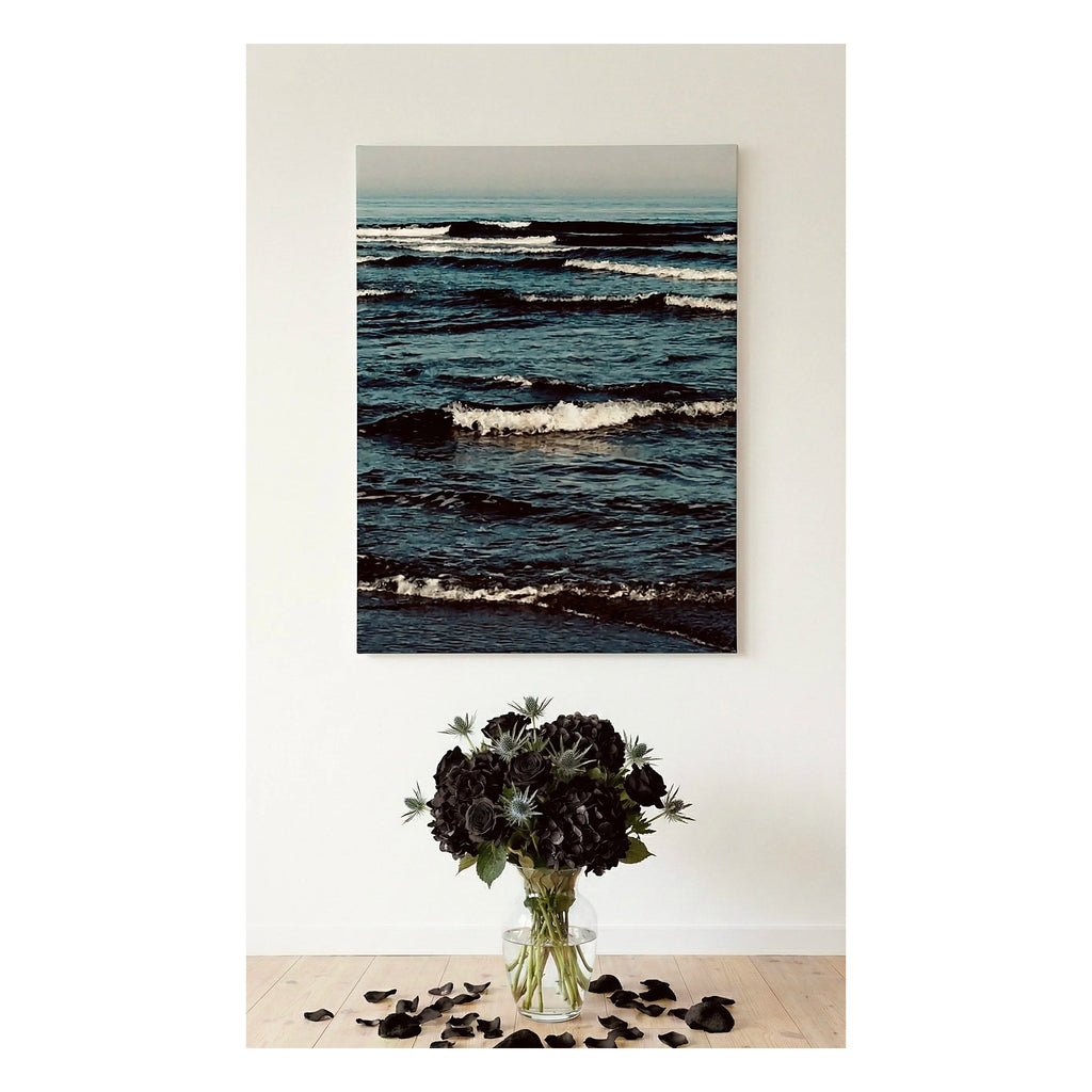 A vertical  Ocean Artwork canvas print of a dark blue ocean with waves, displayed on a white wall above a vase of black flowers on a light wood floor.