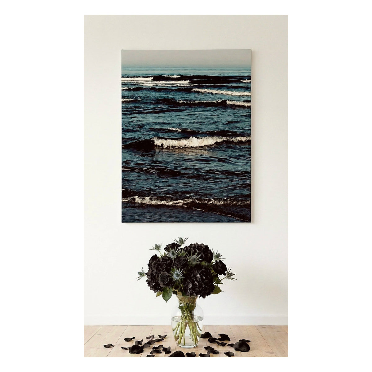 A vertical  Ocean Artwork canvas print of a dark blue ocean with waves, displayed on a white wall above a vase of black flowers on a light wood floor.